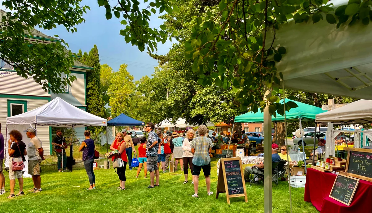 People gathering around food stalls under leafy trees at an outdoor community market.